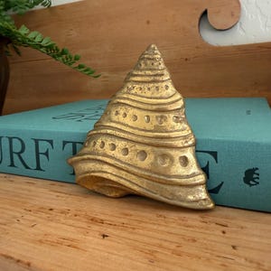 May include: A gold-toned brass seashell figurine with a conical shape, featuring horizontal ridges and a series of small circular indentations along its surface. The object has a weathered, metallic finish and a pointed top, resting in a fixed position.