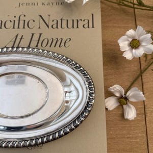 May include: An oval silver tray with a polished, reflective surface and a decorative rope-style border. The tray is positioned on a book titled Pacific Natural At Home, with two white flowers with yellow centers placed nearby on the surface.