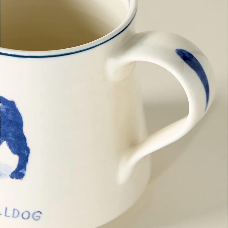 以下が含まれることがあります： 青いブルドッグのイラストと「LDOG」の文字が入った白いセラミックマグ。 マグカップには、青いストライプと縁に青い線が入った湾曲したハンドルが付いています。