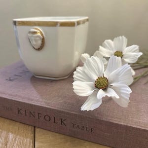 May include: A white ceramic cup with a gold rim and a small gold-toned relief of a face on the side. The cup is placed next to two white artificial cosmos flowers with yellow-green centers, resting on a book titled The Kinfolk Table.