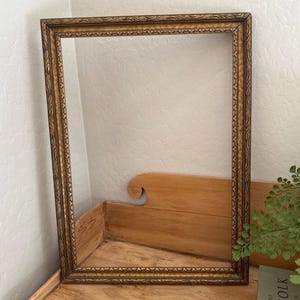 May include: A rectangular, ornate gold picture frame with detailed scrollwork along the edges. The frame is empty, showcasing the intricate design and warm, antique gold color. The frame is resting on a wooden surface.