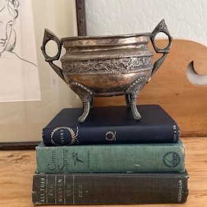 May include: A silver-toned metal bowl with ornate detailing and handles, resting atop a stack of three antique books. The books are in shades of blue, green, and gray, with gold lettering. The bowl has three decorative feet.