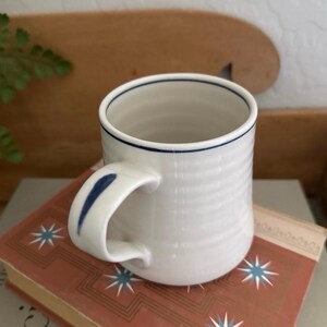 以下が含まれることがあります： 青い縁とハンドルのアクセントが付いた白いセラミックマグ。 マグカップはリブ編みのテクスチャで、星のデザインが施されたヴィンテージの本の上に置かれています。 日常使いに最適なシンプルでエレガントなデザインです。