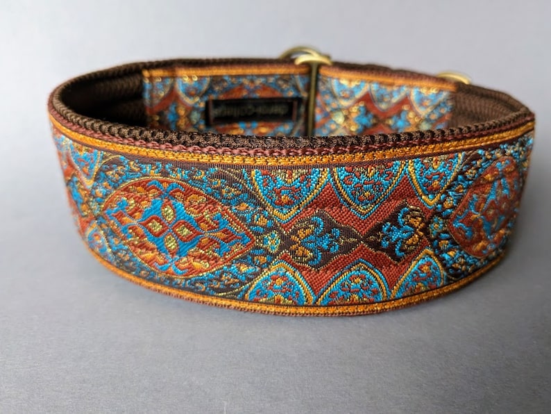 Pu&ograve; includere: Un collare decorativo per cani con un motivo dettagliato nei toni del marrone, verde acqua e oro. Il collare presenta un design tessuto con intricati elementi floreali e geometrici. L'hardware &egrave; color oro.
