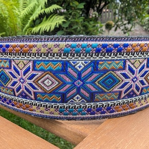 Può includere: Primo piano di una fascia decorativa tessuta con un motivo dettagliato. La fascia presenta disegni geometrici e floreali in blu, rosa, arancione e oro. Probabilmente viene utilizzata per rifinire o abbellire.