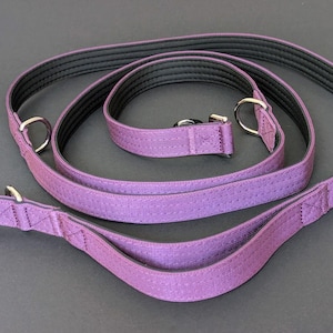 Op de afbeelding: Een paarse hondenriem en bijpassende halsbandset. De riem heeft een zilverkleurige sluiting en een handvat. De halsband heeft een zilveren ring. De set is gemaakt van een textuurmateriaal met een zwart interieur en het merk "derol-collars".