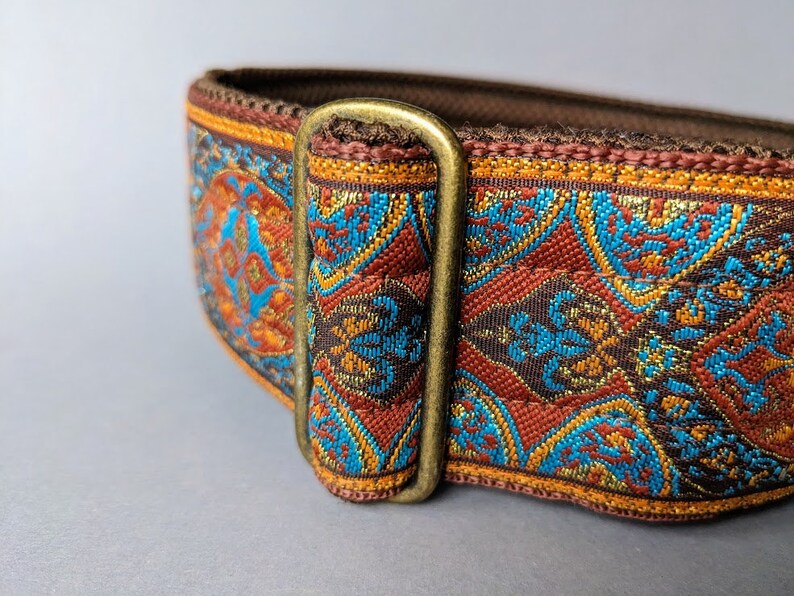 Pu&ograve; includere: Primo piano di un collare per cani con un design ornato e tessuto. Il collare presenta un motivo di forme geometriche turchesi, arancioni e marroni. &Egrave; visibile una fibbia in metallo color ottone. Il collare &egrave; probabilmente per un cane di taglia media o grande.