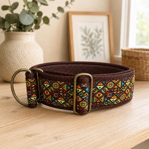 May include: A brown fabric collar featuring a vibrant, multi-colored geometric pattern of triangles and diamonds. The collar includes antique brass-toned metal hardware, specifically two D-rings and a sliding adjuster, with a dark brown mesh lining.