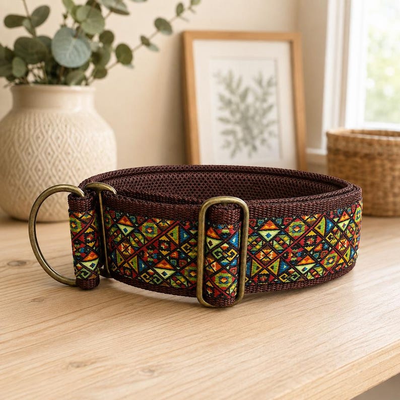 May include: A brown fabric collar featuring a vibrant, multi-colored geometric pattern of triangles and diamonds. The collar includes antique brass-toned metal hardware, specifically two D-rings and a sliding adjuster, with a dark brown mesh lining.