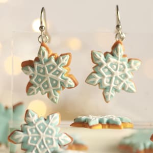 Puede incluir: Pendientes en forma de copo de nieve con ganchos plateados. Los pendientes presentan un diseño de copo de nieve azul claro y blanco con un borde marrón. Se ven otros adornos en forma de copo de nieve. Miden unos 3 cm de diámetro.