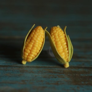 May include: A pair of stud earrings shaped like miniature ears of corn. Each earring features bright yellow kernels arranged in rows, partially encased in light green husks that curve outward at the top, creating a realistic vegetable design.