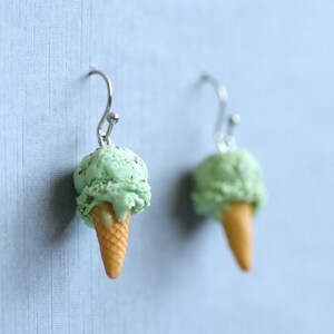 Puede incluir: Un par de pendientes originales con forma de cucuruchos de helado. Cada pendiente tiene una bola de helado verde menta con chispas de chocolate sobre un cono de galleta beige, suspendido de un gancho plateado.