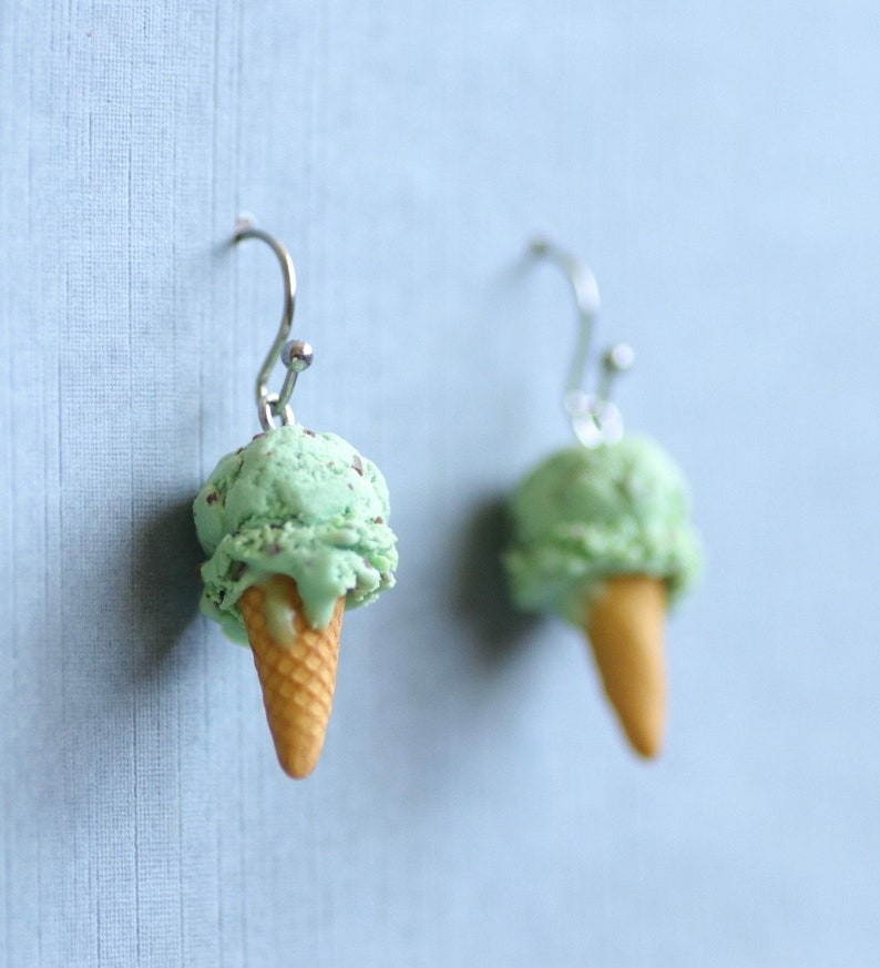 Puede incluir: Un par de pendientes originales con forma de cucuruchos de helado. Cada pendiente tiene una bola de helado verde menta con chispas de chocolate sobre un cono de galleta beige, suspendido de un gancho plateado.
