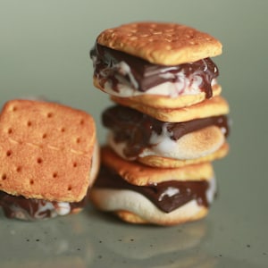 Puede incluir: Una pila de tres s'mores y un s'more individual colocado a la izquierda. Cada s'more consta de dos galletas cuadradas de color marrón dorado, con una capa de chocolate negro derretido y un relleno de malvavisco blanco tostado.