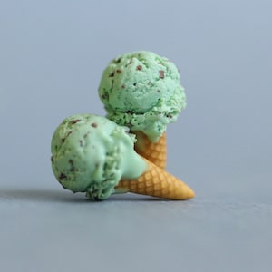 Puede incluir: Dos bolas de helado de menta con chispas de chocolate sobre conos de galleta. El helado es de color verde claro con chispas de chocolate oscuro. Los conos son de color marr&oacute;n claro. El fondo es gris claro liso.