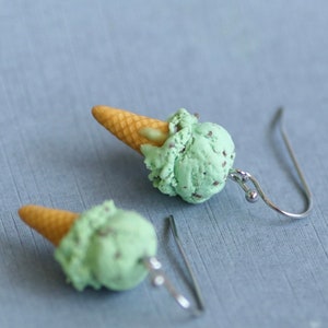 Puede incluir: Un par de pendientes originales con forma de conos de helado en miniatura. Cada pendiente tiene un cono marr&oacute;n claro rematado con una bola de helado verde menta con motas oscuras. Los pendientes tienen ganchos para la oreja de color plateado.