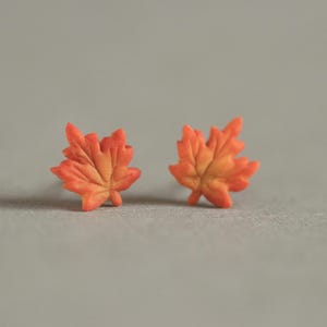 May include: A pair of orange and yellow maple leaf stud earrings. The earrings are made of a material that mimics the look of autumn leaves. The earrings are a fall-themed accessory.