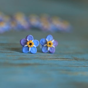 May include: A pair of handcrafted flower stud earrings. Each earring features a blue flower with purple tips, a yellow center, and white accents. The earrings are set against a blue wooden surface, with blurred flowers in the background.