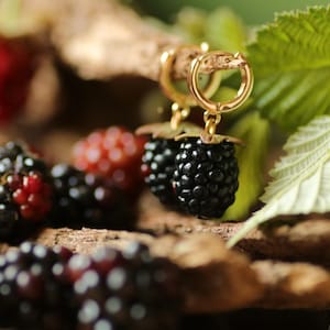 May include: Gold-toned hoop earrings with a blackberry design. The earrings feature a detailed blackberry pendant with a gold-colored leaf and stem. The image also shows fresh blackberries and green leaves.
