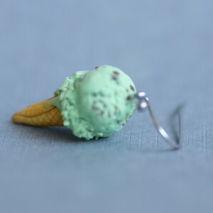 Puede incluir: Un pendiente individual de cono de helado de menta con chispas de chocolate. El helado es verde claro con motas marrones, sobre un cono de gofre beige. El pendiente tiene un gancho plateado.