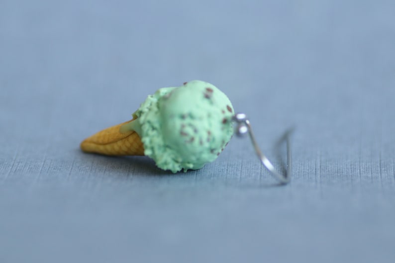 Puede incluir: Un pendiente individual de cono de helado de menta con chispas de chocolate. El helado es verde claro con motas marrones, sobre un cono de gofre beige. El pendiente tiene un gancho plateado.