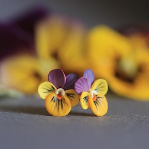 May include: Close-up of two pansy flowers with yellow petals, purple and yellow accents, and a dark center. The background features blurred yellow and purple flowers, creating a soft focus effect. The flowers are on a gray surface.