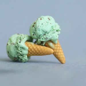 Puede incluir: Dos conos de helado de menta con chispas de chocolate, con bolas de color verde claro y chispas de chocolate marr&oacute;n. Los conos son de color marr&oacute;n claro y descansan sobre una superficie gris.