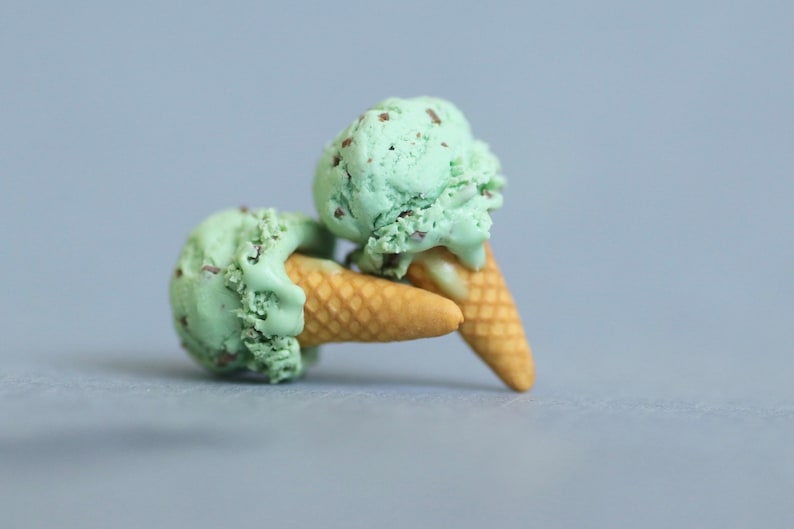 Puede incluir: Dos conos de helado de menta con chispas de chocolate, con bolas de color verde claro y chispas de chocolate marr&oacute;n. Los conos son de color marr&oacute;n claro y descansan sobre una superficie gris.