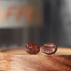 May include: Two roasted coffee beans sit on a small, round wooden tray. The beans are a rich, dark brown color, with visible textures. The background is blurred, with hints of orange text.