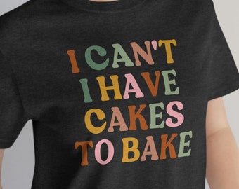 Puede incluir: Una camiseta negra con el texto "I CAN'T I HAVE CAKES TO BAKE" en letras de colores.