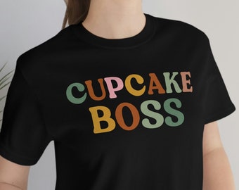Puede incluir: Camiseta negra con el texto "CUPCAKE BOSS" en letras de colores.