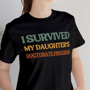 May include: A black t-shirt with the text "I survived my daughter's doctorate program" in green, orange, and yellow.