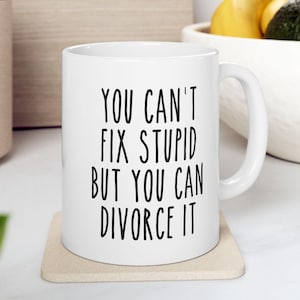 May include: White ceramic mug with the text "YOU CAN'T FIX STUPID BUT YOU CAN DIVORCE IT" in black. The mug is on a beige coaster. The background includes a wooden surface and a bowl with fruit.