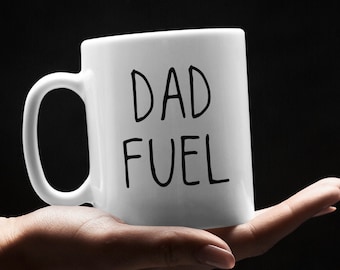 May include: White ceramic mug with the words "DAD FUEL" in black lettering. The mug is held in a person's hand against a black background. The mug has a curved handle.