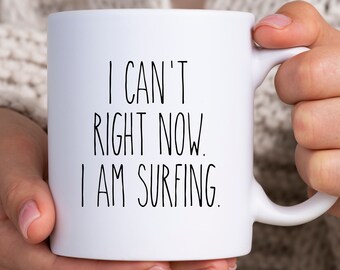 Könnte beinhalten: Weiße Keramik-Tasse mit dem Text "I CAN'T RIGHT NOW. I AM SURFING." in schwarzer, handschriftlicher Schrift. Die Tasse wird von einer Person gehalten. Die Tasse hat eine Standardgröße für Kaffee oder Tee.