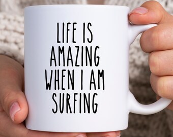 Könnte beinhalten: Weißer Keramikbecher mit dem Text "LIFE IS AMAZING WHEN I AM SURFING" in schwarzen, fetten Buchstaben. Der Becher hat einen Standardgriff und wird von einer Person gehalten.