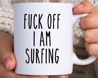 Könnte beinhalten: Weiße Keramik-Tasse mit dem Text "FUCK OFF I AM SURFING" in schwarzen, fetten Buchstaben. Die Tasse hat einen Standardgriff und wird von einer Person gehalten.