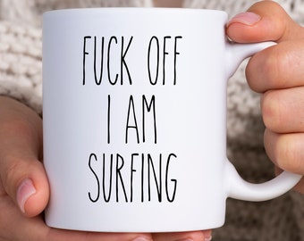 Könnte beinhalten: Weiße Keramik-Tasse mit dem Text "FUCK OFF I AM SURFING" in schwarzer, handschriftlicher Schrift. Die Tasse hat einen Standardgriff und wird von einer Person gehalten.