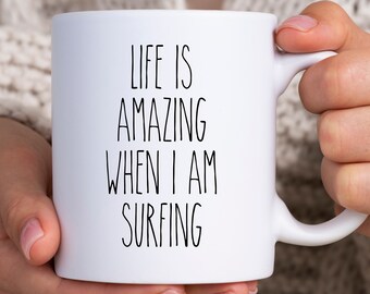 Könnte beinhalten: Weiße Keramik-Tasse mit dem Text "LIFE IS AMAZING WHEN I AM SURFING" in Schwarz. Die Tasse hat einen gebogenen Henkel und wird von einer Person gehalten.