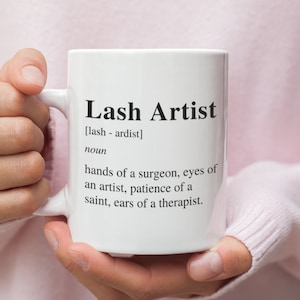 以下が含まれることがあります： 黒いデザインで「Lash Artist [lash - ardist] noun hands of a surgeon, eyes of an artist, patience of a saint, ears of a therapist.」と書かれた白いセラミックマグカップ