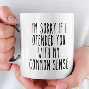 以下が含まれることがあります： 「I'M SORRY IF I OFFENDED YOU WITH MY COMMON SENSE」のテキストが黒いブロック体で書かれた白いセラミックマグ。マグカップには灰色のハンドルがあり、人が持っています。