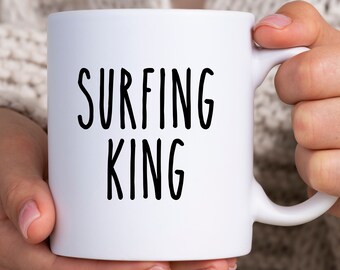 May include: White ceramic mug with the text "SURFING KING" in black, bold letters. The mug is held by a person. The mug has a standard shape and a curved handle.