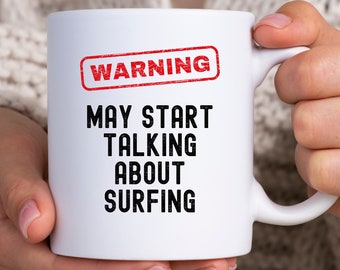 Könnte beinhalten: Weiße Keramik-Tasse mit dem Text "WARNING MAY START TALKING ABOUT SURFING" in Schwarz, mit einem roten "WARNING"-Stempel. Die Tasse wird von einer Person gehalten.