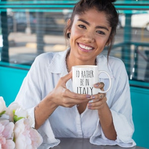 May include: A white ceramic mug with black text that reads "I'd rather be in Italy". The mug is being held by a person with a white shirt and a bun.