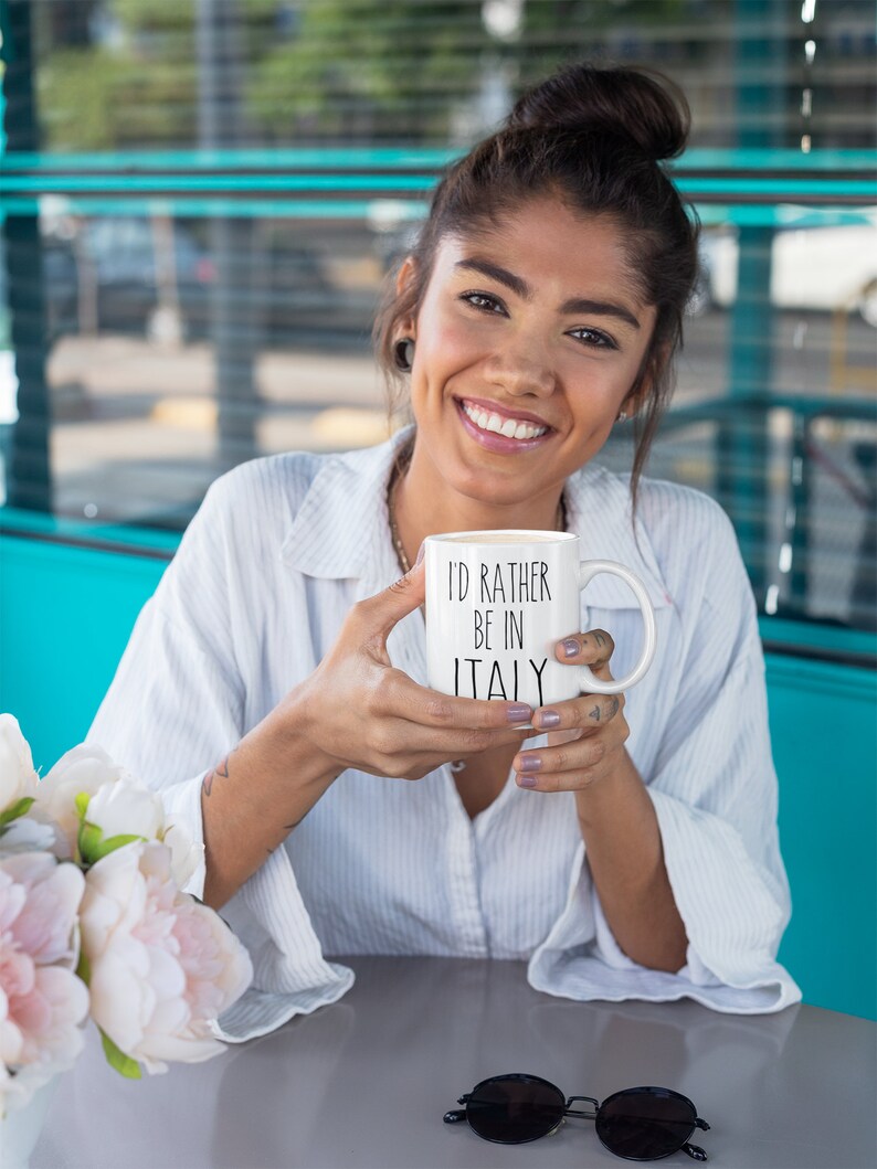 May include: A white ceramic mug with black text that reads "I'd rather be in Italy". The mug is being held by a person with a white shirt and a bun.