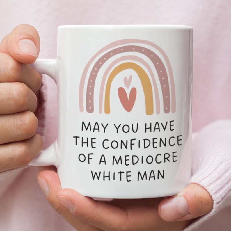 May include: White ceramic mug with a handle, featuring a rainbow design in shades of pink, peach, and gold. The mug has the text "MAY YOU HAVE THE CONFIDENCE OF A MEDIOCRE WHITE MAN" printed in black. The mug is held by a person.