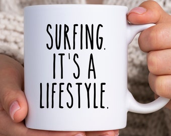 Könnte beinhalten: Weiße Keramik-Tasse mit dem Text "SURFING. IT'S A LIFESTYLE" in schwarzer, fetter, handschriftlicher Schrift. Die Tasse wird von einer Person gehalten. Die Tasse hat einen Standardgriff.