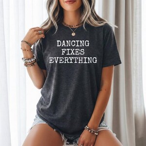 Könnte beinhalten: Dunkelgraues T-Shirt mit dem weißen Text "DANCING FIXES EVERYTHING". Das T-Shirt hat einen Rundhalsausschnitt und scheint aus einem weichen Material zu bestehen. Der Stil ist lässig und bequem.
