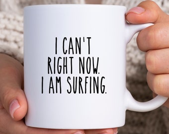 Könnte beinhalten: Weiße Keramik-Tasse mit dem Text "I CAN'T RIGHT NOW. I AM SURFING" in schwarzer, serifenloser Schrift. Die Tasse hat einen gebogenen Henkel und wird von einer Person gehalten.