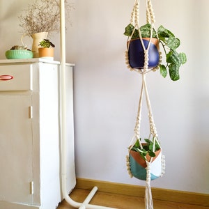 Puede incluir: Dos maceteros colgantes de macramé con cuerda de algodón blanca y cuentas de madera. El macetero superior tiene un tiesto azul oscuro con una planta verde. El macetero inferior tiene un tiesto azul, blanco y marrón con una planta verde.